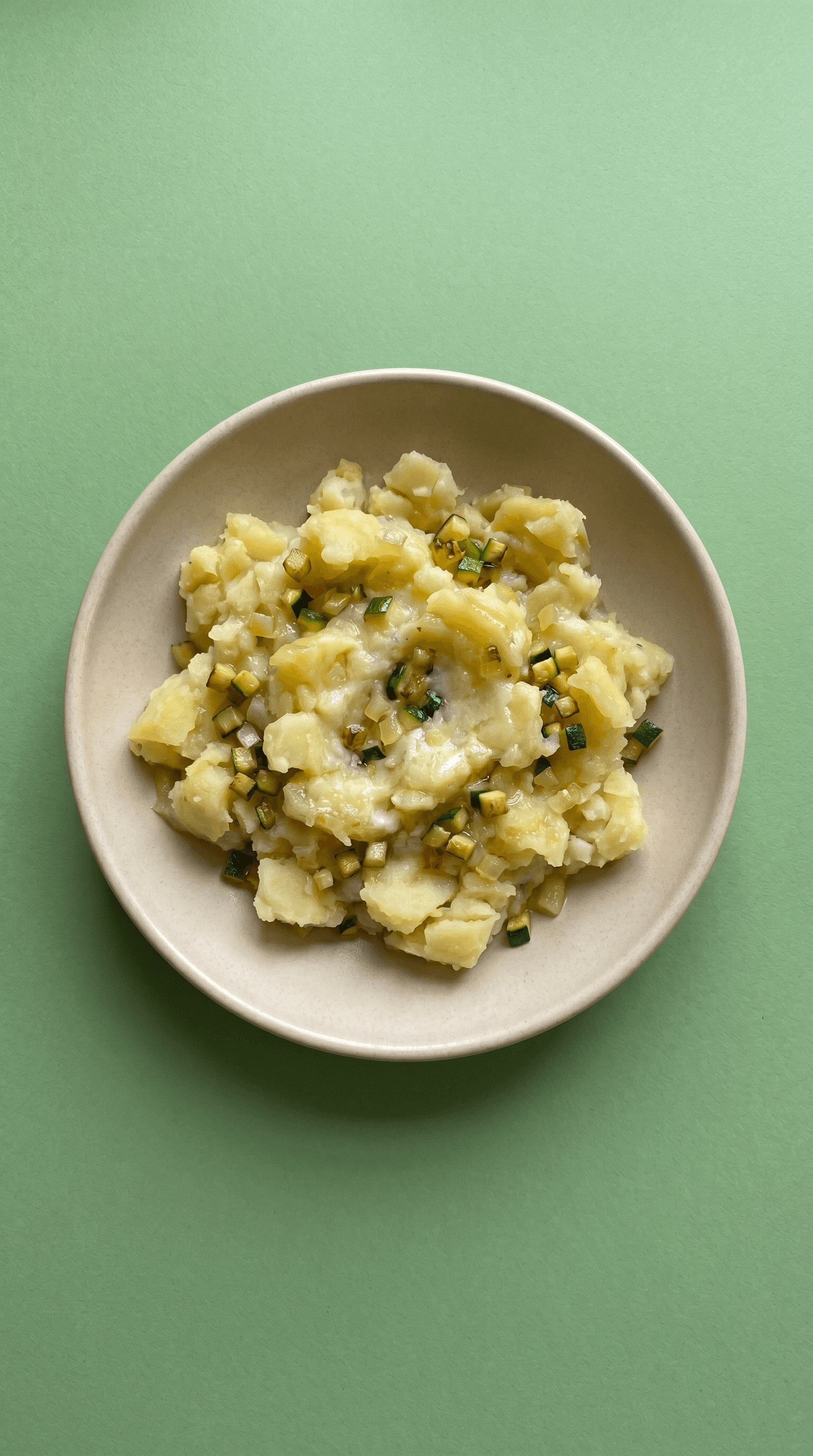 Écrasé de pommes de terre aux courgettes et citron sur une assiette beige sur un fond vert sauge, un accompagnement rustique prêt à réchauffer et servir.