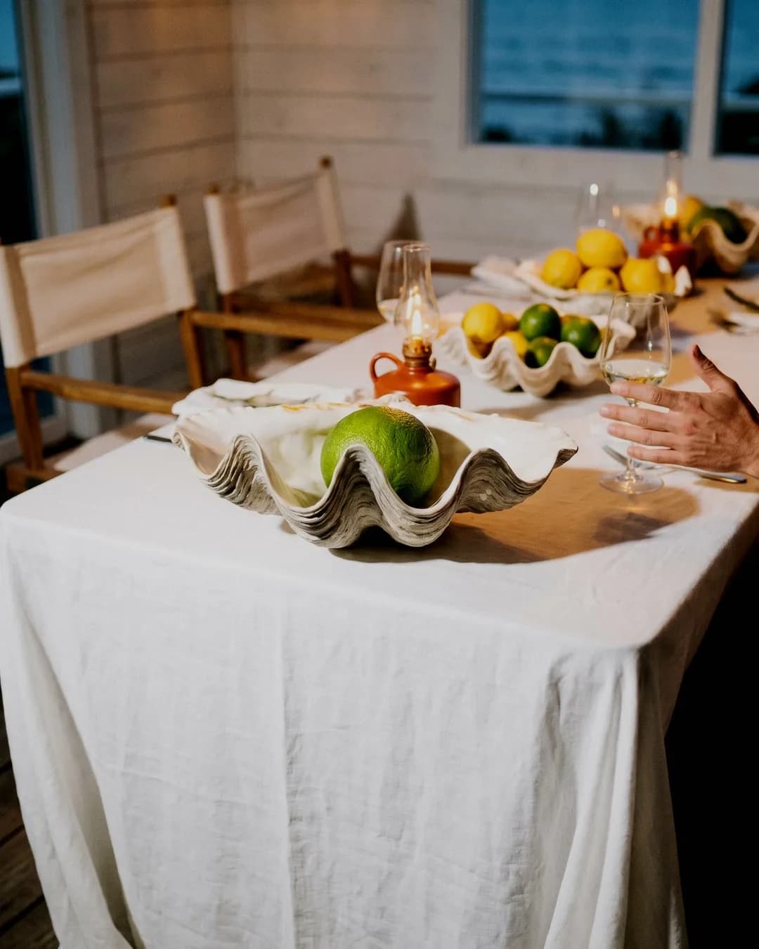 Witte linnen tafel met schelpvormige schaal, groene citrus, kaarslicht en wijnglazen in een intieme setting