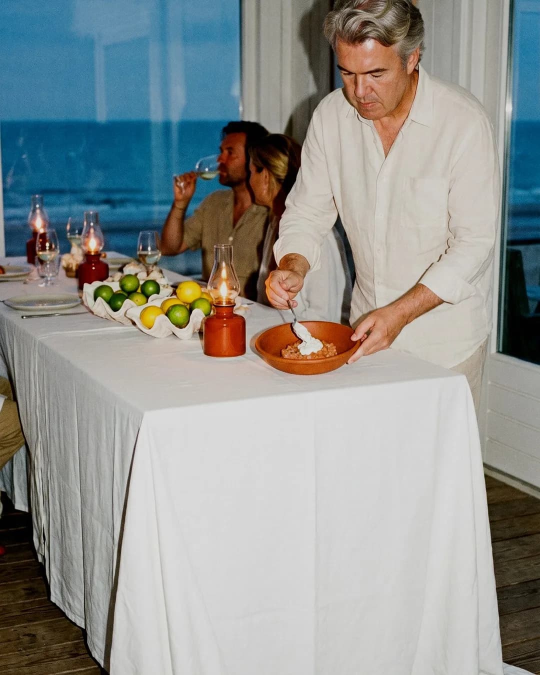 Le chef Christophe Rammant de Mr. Saucy finalise un plat sur une table dressée avec vue sur la mer, tandis que des invités dégustent du vin en arrière-plan.