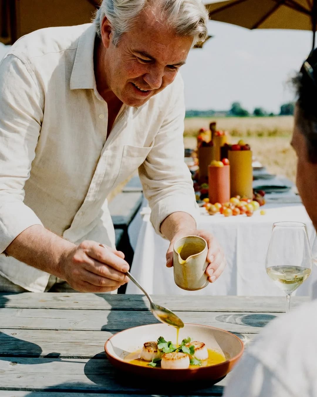 Le chef Christophe Rammant verse une sauce sur une assiette de noix de Saint-Jacques poêlées, tout en souriant à un invité attablé autour d'une table en bois.