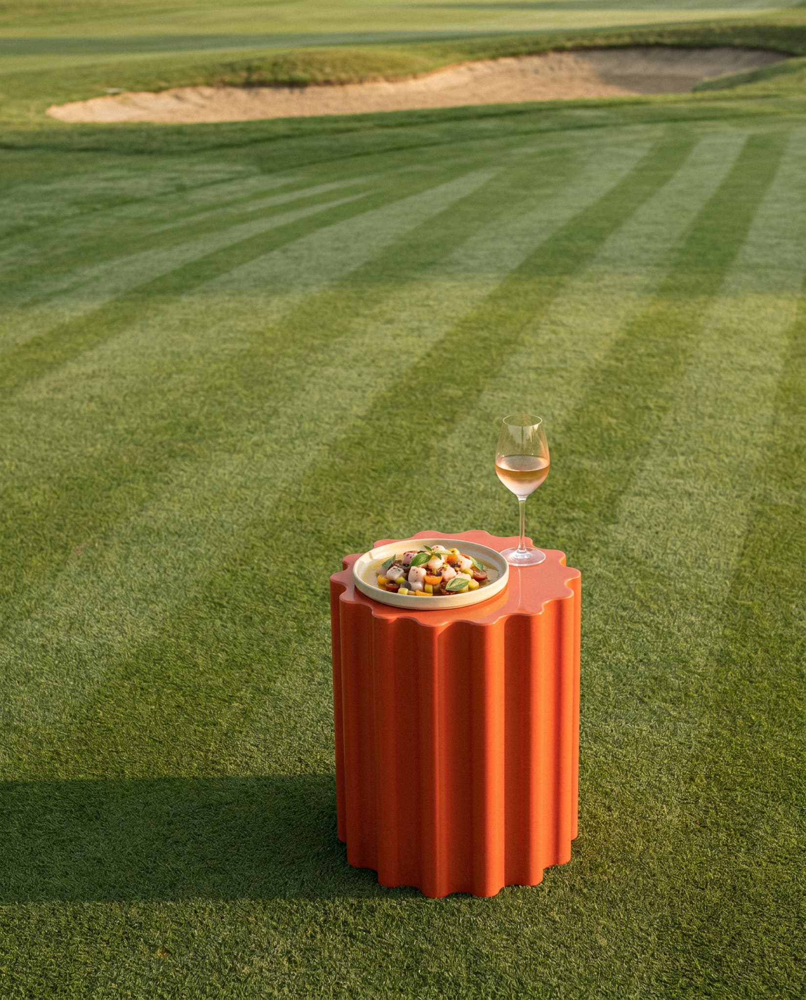 Une table élégante aux tons corail et orange sur un fairway de golf impeccable, dressée avec une assiette de fruits de mer saisis et un verre de vin blanc