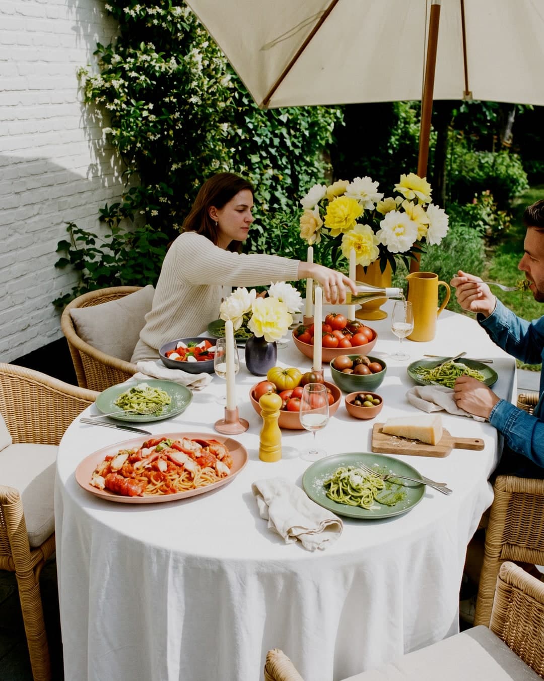Table dressée au soleil sur une plage, nappée de blanc, avec un poisson entier grillé, des pommes de terre, des tomates et des convives partageant un repas