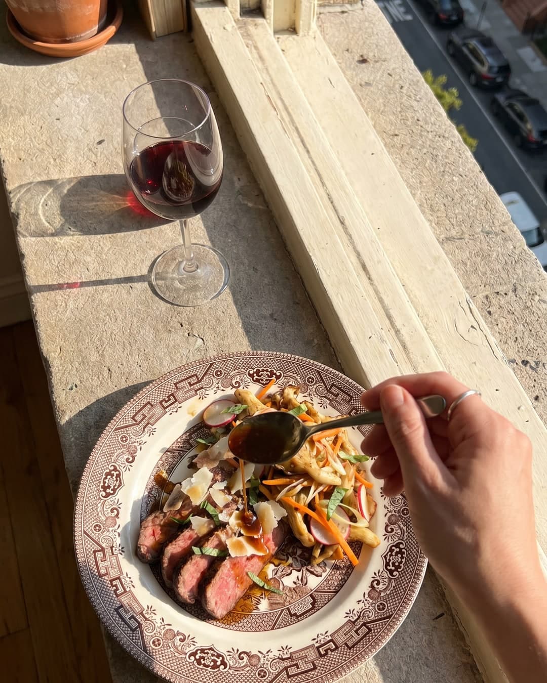 Un cliché pris sur le vif, en vue plongeante, d'une assiette en céramique à motifs brun et crème posée sur un rebord de fenêtre en pierre baigné de soleil, accompagnée d'un verre de vin rouge et d'une main versant une sauce sur du bœuf émincé et des champignons.