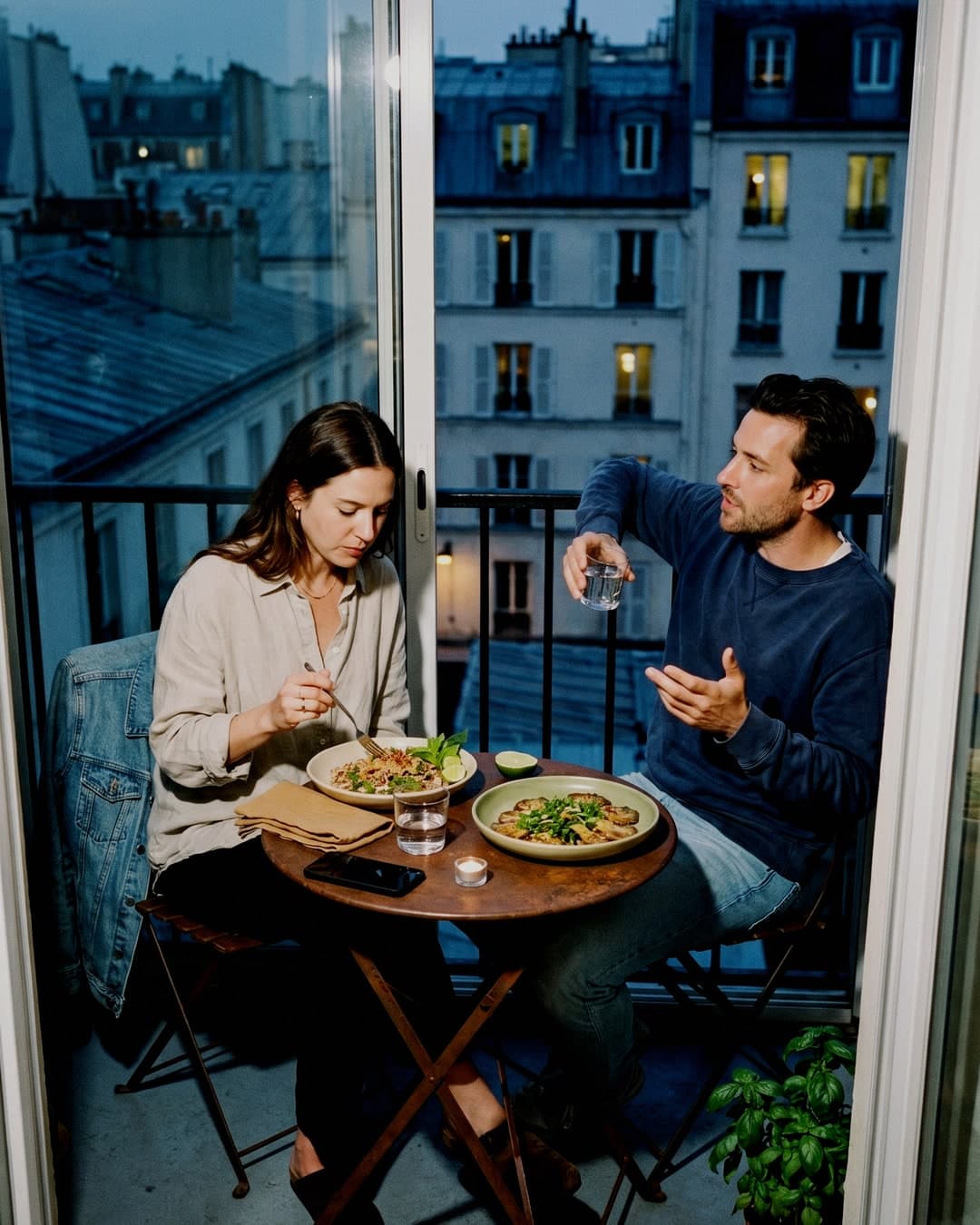 Couple enjoying dinner on a small balcony at blue hour with wine and warm interior light